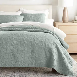 A bedroom with a pastel green quilt featuring a swirling pattern, two matching pillows, and a light wood dresser with a ceramic lamp.