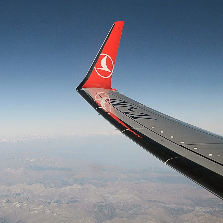 Airplane wing with a red Turkish Airlines winglet and logo, flying over mountainous terrain under a clear blue sky.