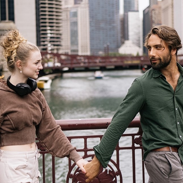A woman wearing a brown cropped hoodie and headphones holds hands with a man in a green button-up shirt on a bridge with a cityscape in the background.