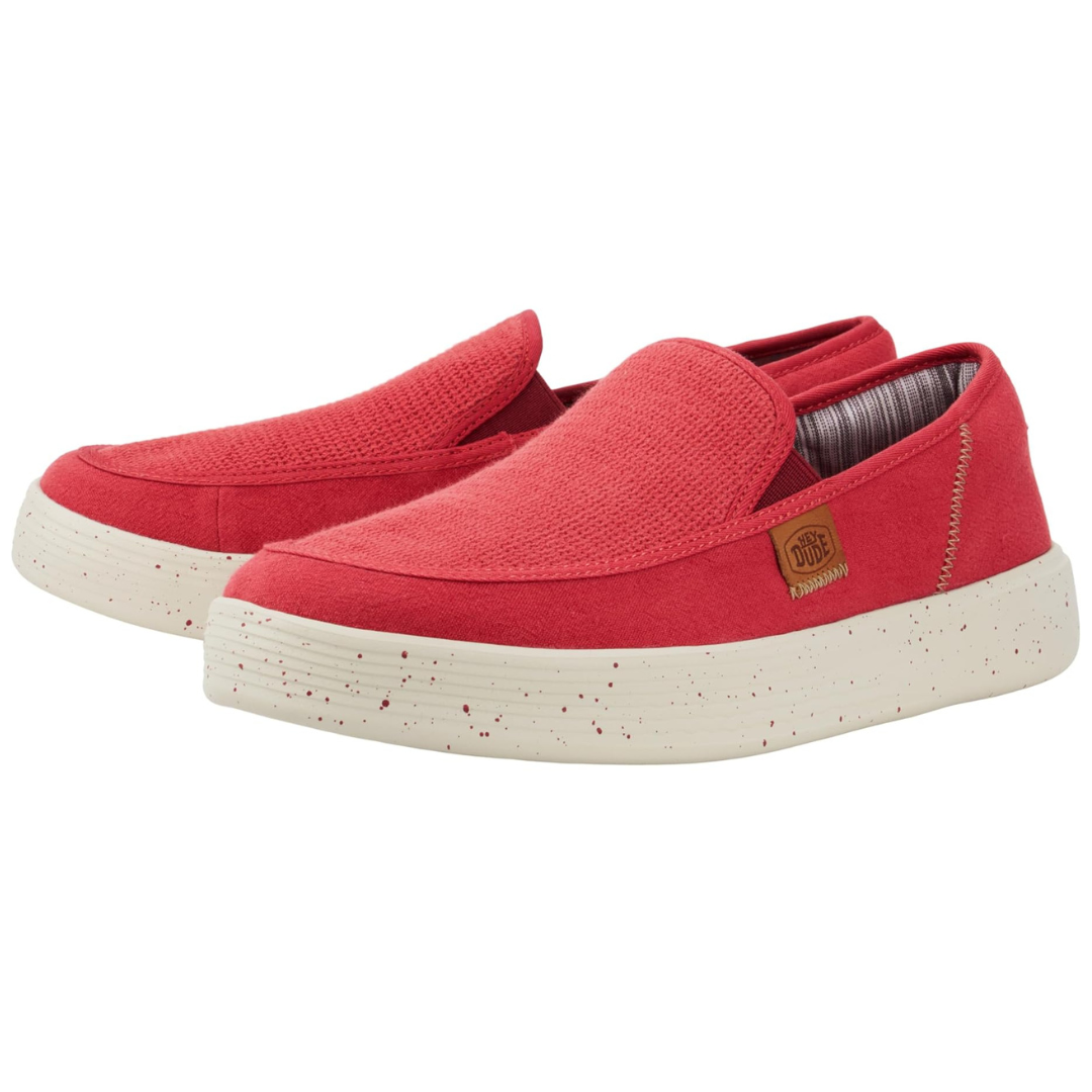 Red slip-on shoes with a textured upper, speckled white soles, and a small brown \“Hey Dude\“ label on the side. They feature a casual and relaxed style, suitable for everyday wear.