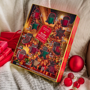 Holiday-themed candle advent calendar with 12 scented candles, featuring fragrances like Christmas Wreath, Hot Apple Cider, and Spiced Gingerbread. Decorated with festive red and green shapes, surrounded by red ornaments and gifts.