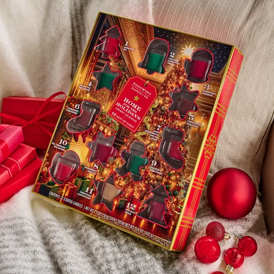 Holiday-themed candle advent calendar with 12 scented candles, featuring fragrances like Christmas Wreath, Hot Apple Cider, and Spiced Gingerbread. Decorated with festive red and green shapes, surrounded by red ornaments and gifts.