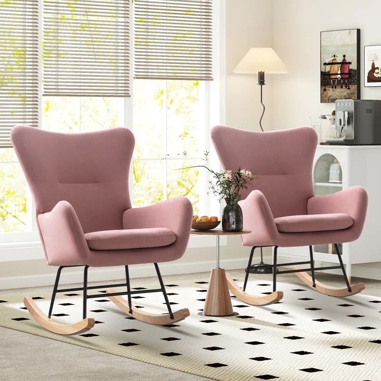 Two pink upholstered rocking chairs with wooden runners are positioned in a bright room near a window. A small round table with a vase and snacks sits between them on a patterned rug.