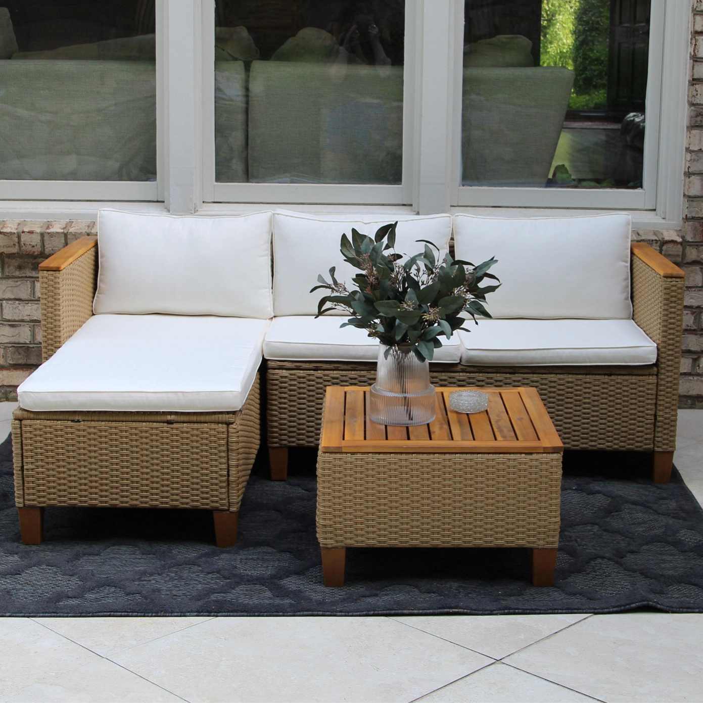 Outdoor patio set with white cushions includes a wicker L-shaped sofa, a matching square ottoman, and a wooden-topped coffee table featuring a vase with greenery, all on a dark patterned rug.