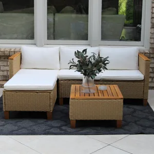 Outdoor patio set with white cushions includes a wicker L-shaped sofa, a matching square ottoman, and a wooden-topped coffee table featuring a vase with greenery, all on a dark patterned rug.