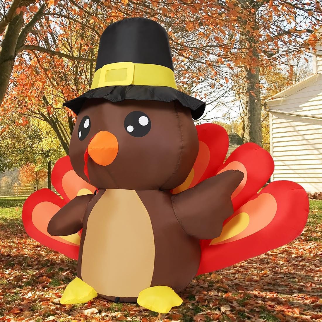 An inflatable turkey wearing a pilgrim hat stands outdoors among autumn leaves, with red and orange tail feathers.