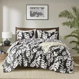 A bed features a black and white floral-patterned comforter set, with matching pillows. The room includes a lamp on a side table, framed abstract art above the bed, and a potted plant on the right.