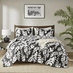 A bed features a black and white floral-patterned comforter set, with matching pillows. The room includes a lamp on a side table, framed abstract art above the bed, and a potted plant on the right.