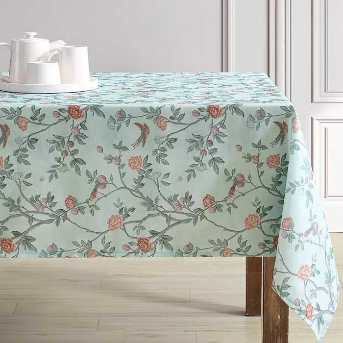Floral tablecloth with a pastel background featuring branches, leaves, and birds in a detailed pattern, paired with a white teapot and cups on a tray.