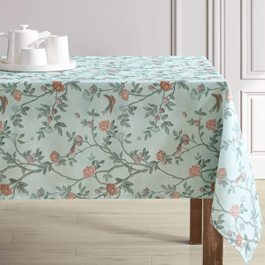 Floral tablecloth with a pastel background featuring branches, leaves, and birds in a detailed pattern, paired with a white teapot and cups on a tray.