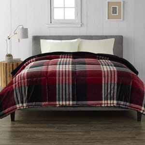 Cuddl Duds Comforter Set features a red, black, and gray plaid pattern, with a plush design for added comfort, shown with light yellow pillows and a gray upholstered headboard. The setting includes a minimalist nightstand with a lamp and decor.