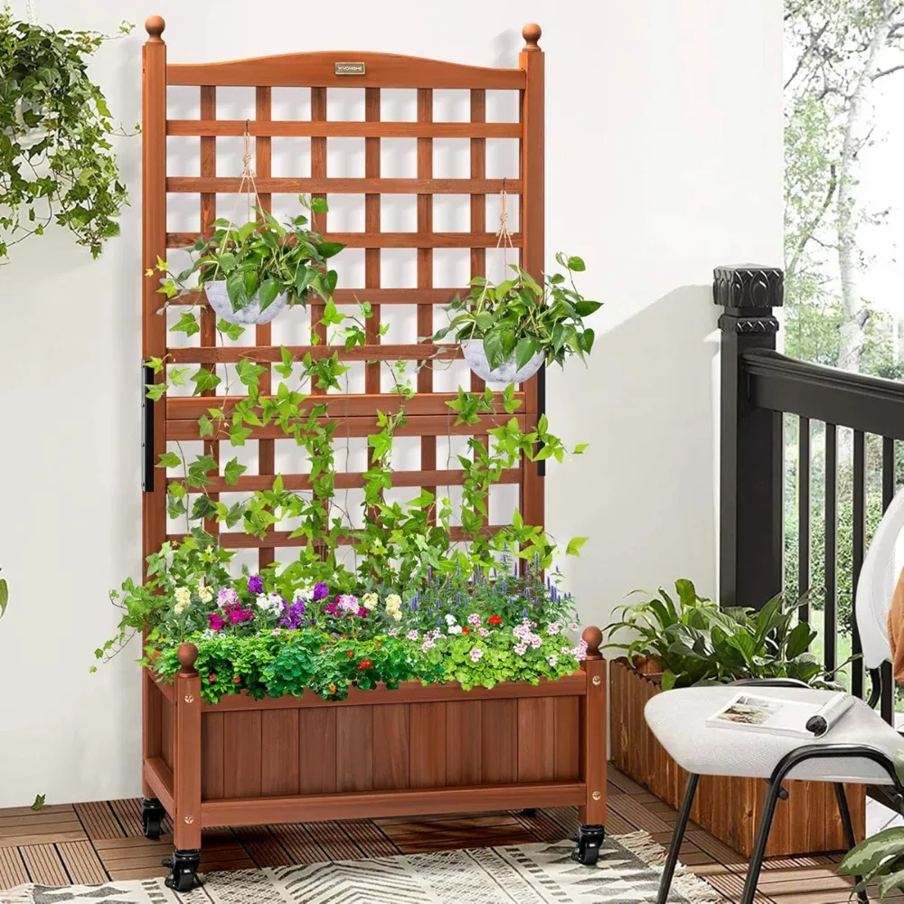 A wooden planter box with an attached trellis supports climbing plants, with hanging pots and flowers below, placed on a balcony with additional decor.