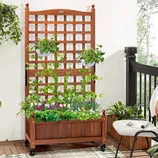 A wooden planter box with an attached trellis supports climbing plants, with hanging pots and flowers below, placed on a balcony with additional decor.
