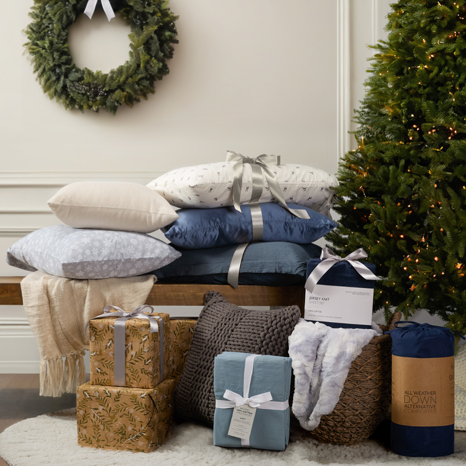 Stacked pillows, cozy blankets, and knit throw pillows are near a decorated Christmas tree. Wrapped gifts are topped with bows, and a festive wreath hangs on the wall. Various bedding items, including a down alternative comforter, are also shown.