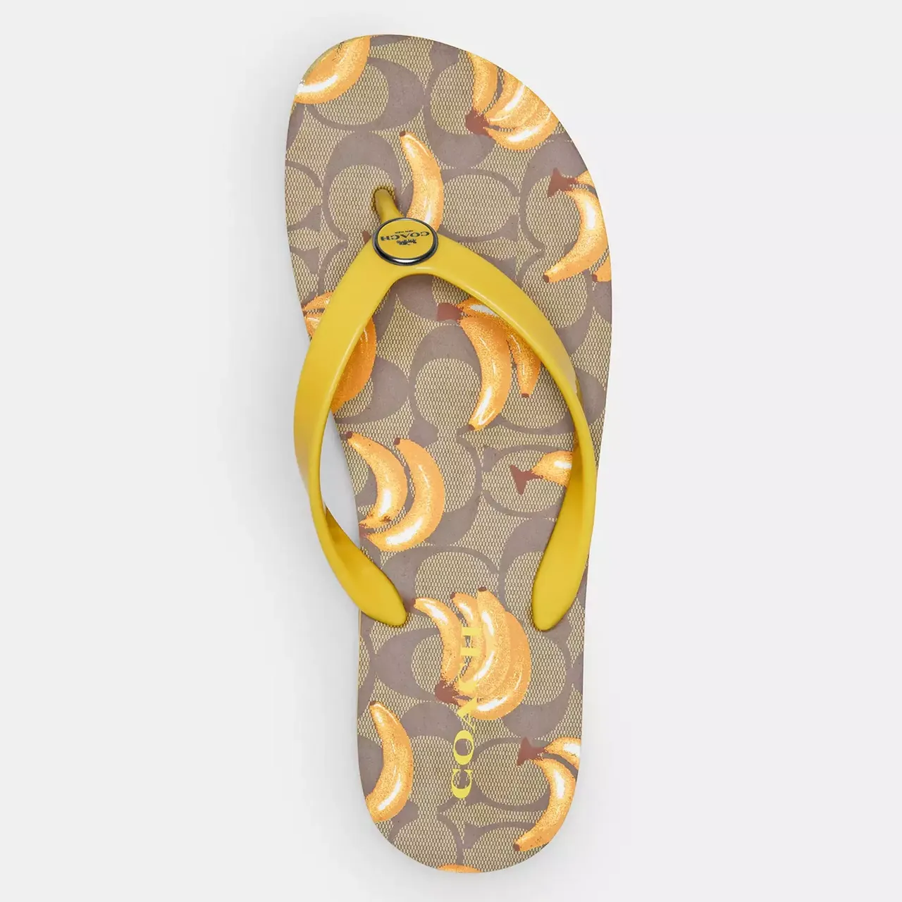 A single sandal with a fruit print featuring sliced bananas on a patterned background, with a yellow thong strap bearing the Coach brand logo.