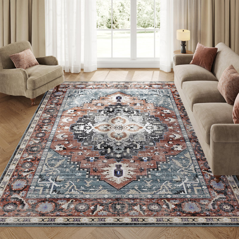 Intricately patterned rug featuring a central geometric medallion design in rust, blue, and cream tones, accented with floral motifs, perfectly complementing a neutral living room setting with beige seating and soft lighting.