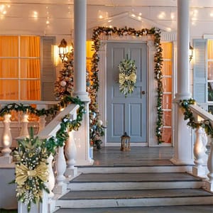 A festive porch with garlands wrapped around columns, door frame, and railings, all adorned with string lights. A decorative wreath hangs on the door, and a lantern is placed at the entrance.