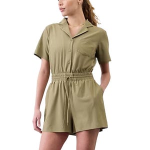 A woman is wearing an olive green short-sleeve jumpsuit with a button-up front, elastic waistband with a drawstring, and a chest pocket.