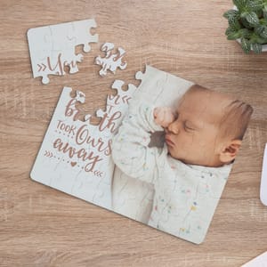 A partially assembled jigsaw puzzle shows a sleeping baby and the text \“Just took ours away\“ in a decorative font. The puzzle is displayed on a wooden surface alongside a small potted plant.