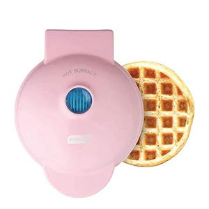 A pink Dash Mini Waffle Maker with a textured surface and a blue heat indicator light is shown, accompanied by a round, golden-brown waffle partially visible beneath it.