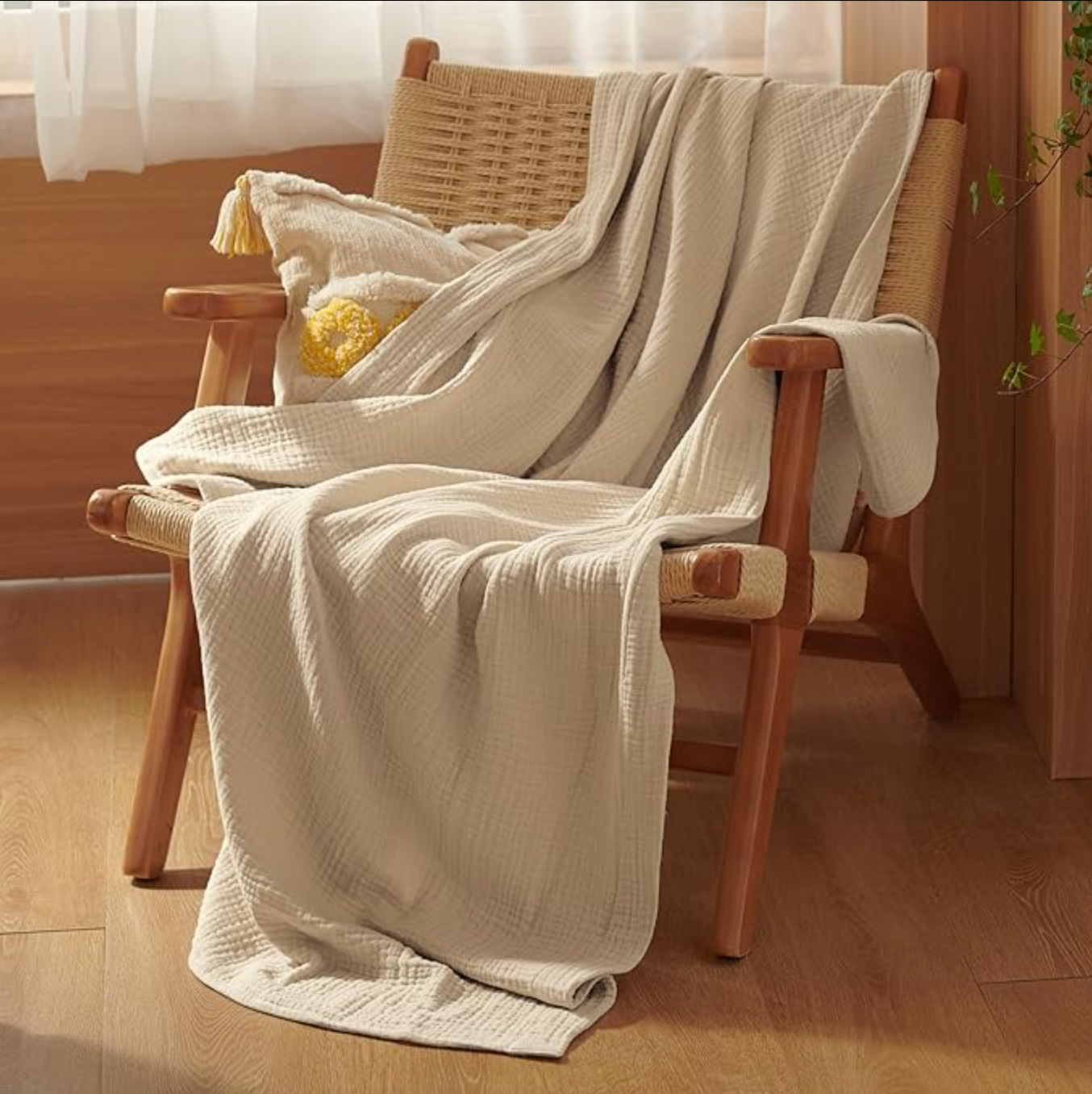 A cream-colored, textured cotton muslin throw blanket drapes over a woven wooden chair, accompanied by a tasseled cushion with a yellow accent.