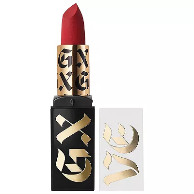 A red matte lipstick with a black case that features bold gold lettering, accompanied by two gold adhesive wing-shaped designs for applying to the skin.