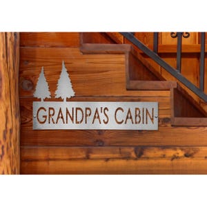 A custom steel sign mounted on wooden stairs, reading \“GRANDPA'S CABIN\“ with two tree silhouettes beside the text, reflecting a rustic cabin theme.