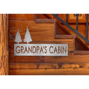 A custom steel sign mounted on wooden stairs, reading \“GRANDPA'S CABIN\“ with two tree silhouettes beside the text, reflecting a rustic cabin theme.