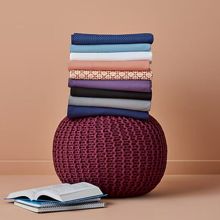 A stack of folded clothes in various colors sits atop a round, burgundy knitted pouf. An open book and a spiral notebook are placed on the floor nearby.