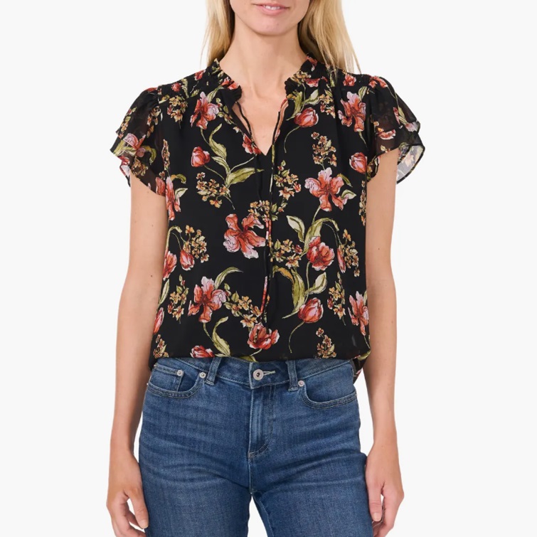 A woman wearing a black floral blouse with ruffled cap sleeves paired with blue jeans.