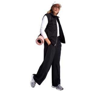 A person wearing a black sleeveless jacket, white long-sleeve shirt, and black pants, with gray sneakers and a white cap, carries a pink yoga mat.