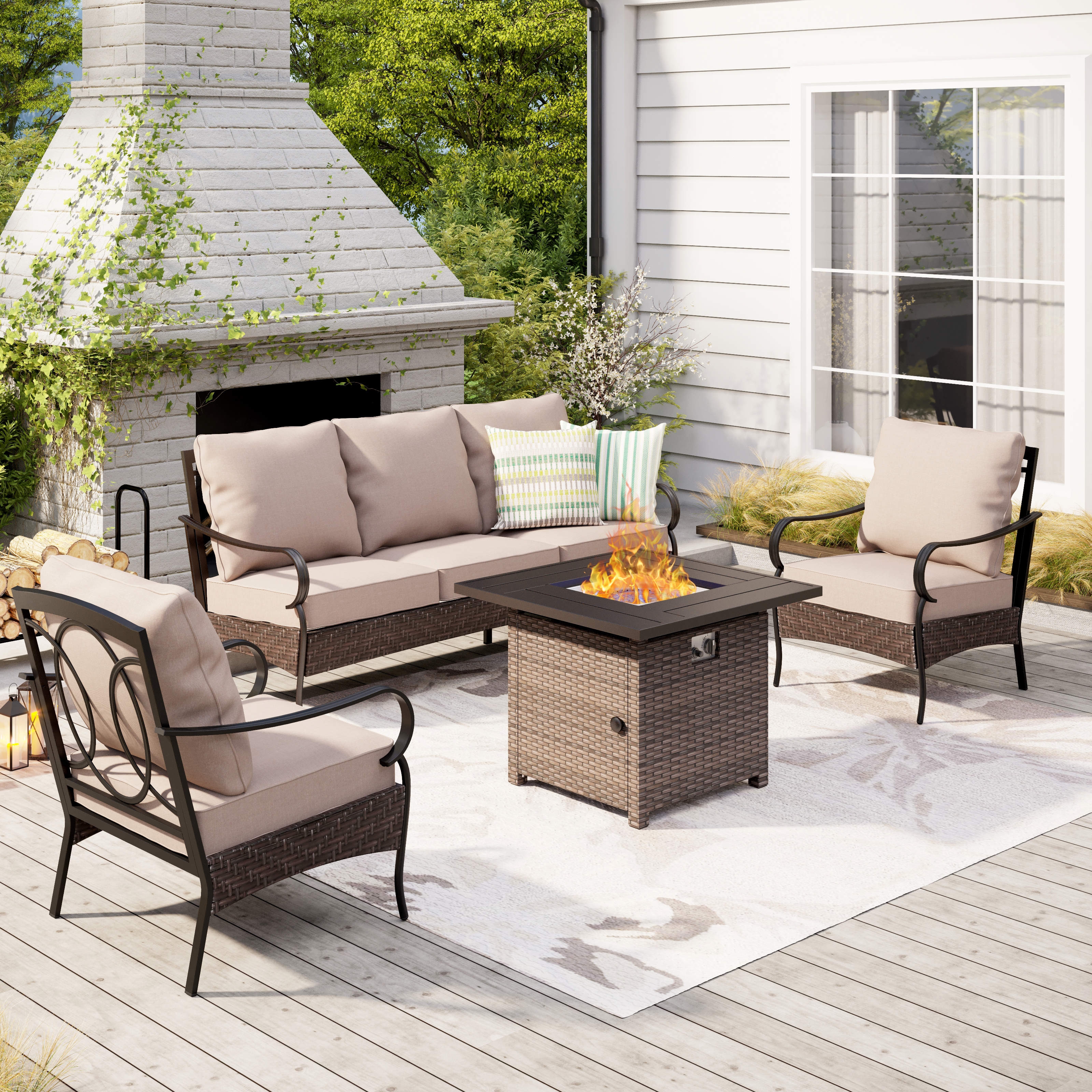Outdoor patio set with beige cushioned sofa and chairs, wicker fire pit table, and decorative pillows on a wooden deck with a brick fireplace backdrop.
