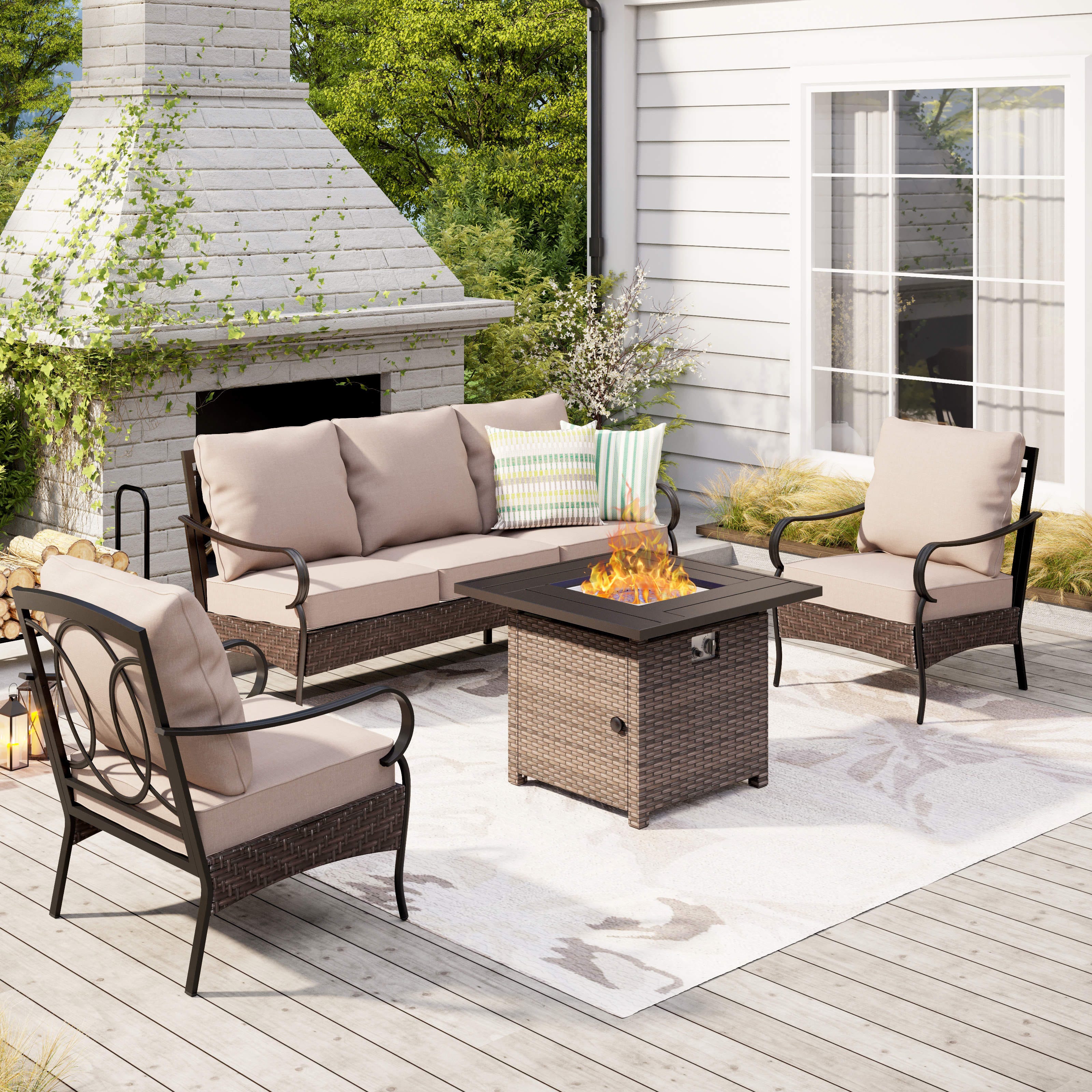 Outdoor patio set with beige cushioned sofa and chairs, wicker fire pit table, and decorative pillows on a wooden deck with a brick fireplace backdrop.