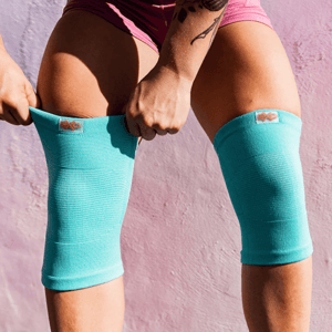 A person is wearing turquoise knee sleeves, slightly pulling them up. The sleeves feature a small logo and are designed for athletic support.