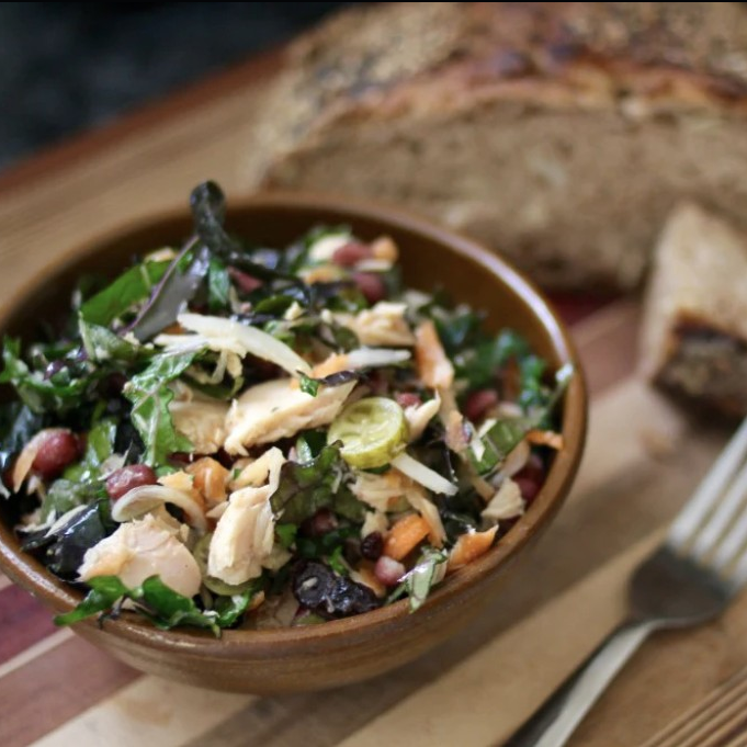 A bowl of mixed salad with leafy greens, shredded vegetables, and chicken pieces is next to a fork and rustic bread on a wooden board.