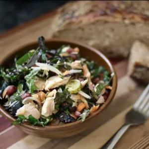 A bowl of mixed salad with leafy greens, shredded vegetables, and chicken pieces is next to a fork and rustic bread on a wooden board.