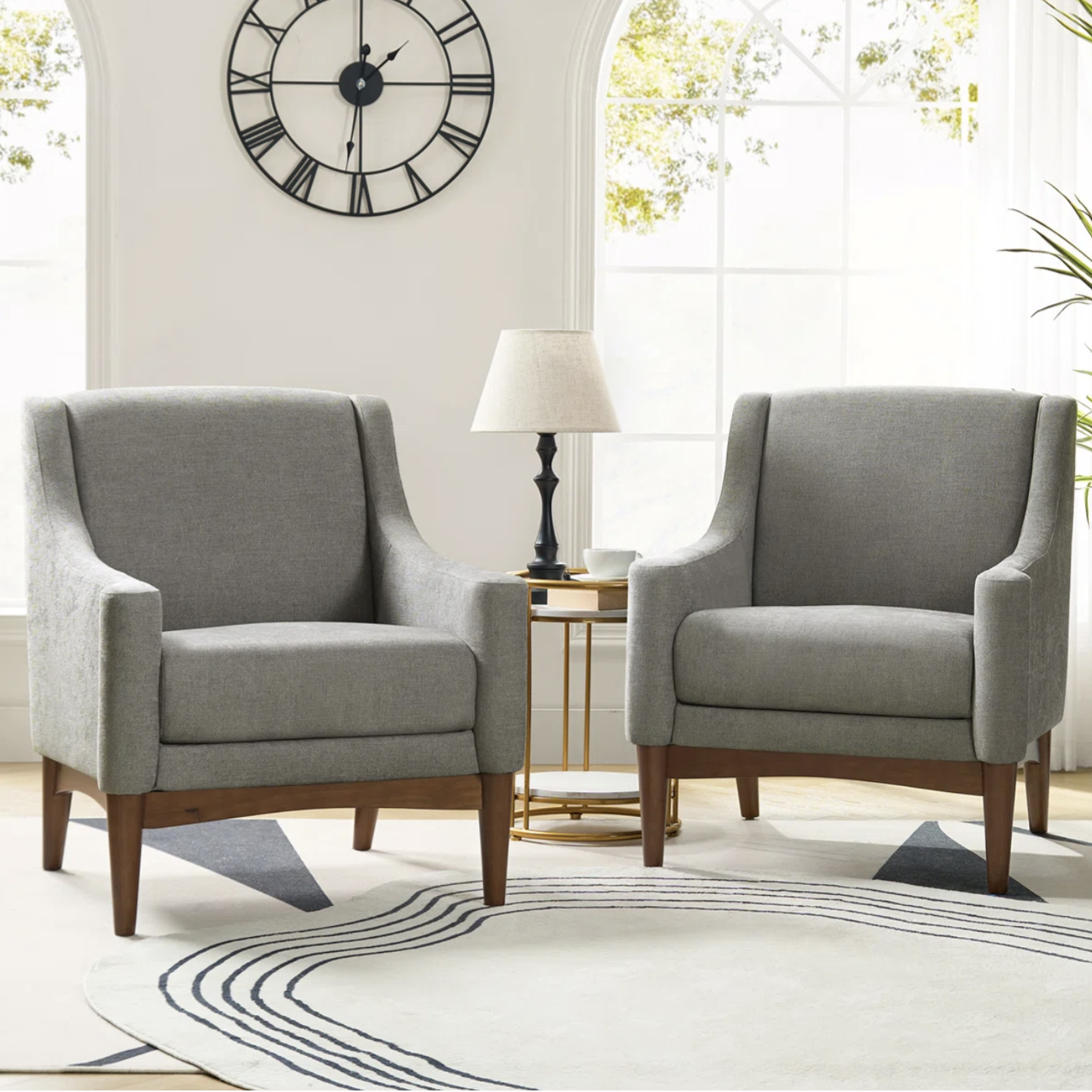 Two gray upholstered armchairs with wooden legs, flanking a small round table with a lamp. Behind them is a large clock on the wall and arched windows, with a patterned rug on the floor.