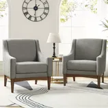 Two gray upholstered armchairs with wooden legs, flanking a small round table with a lamp. Behind them is a large clock on the wall and arched windows, with a patterned rug on the floor.