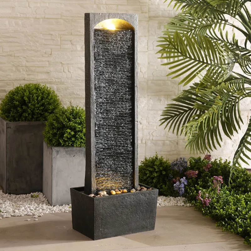 A tall, rectangular water fountain with a textured surface and built-in light, surrounded by decorative plants and pebbles on a patio.