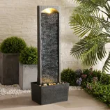 A tall, rectangular water fountain with a textured surface and built-in light, surrounded by decorative plants and pebbles on a patio.