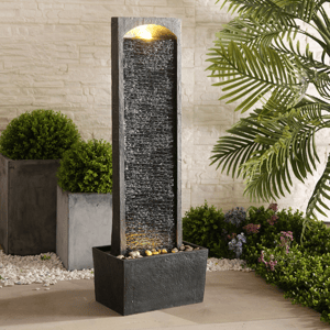 A tall, rectangular water fountain with a textured surface and built-in light, surrounded by decorative plants and pebbles on a patio.