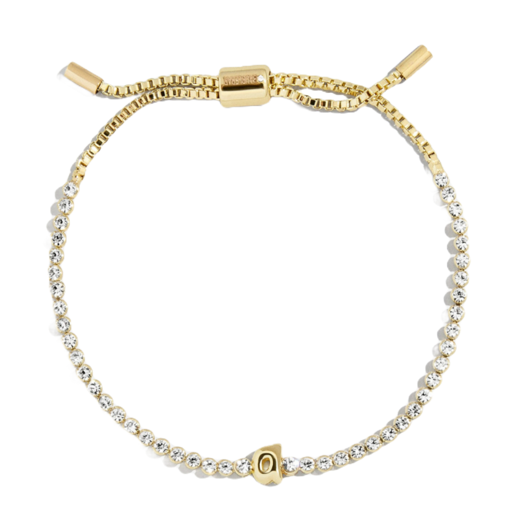 A gold tennis bracelet featuring clear gemstones along its length and a customizable bubble initial charm. It includes an adjustable sliding clasp for fitting.