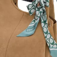 A silk skinny scarf with a green and white Coach signature print tied to a tan handbag.