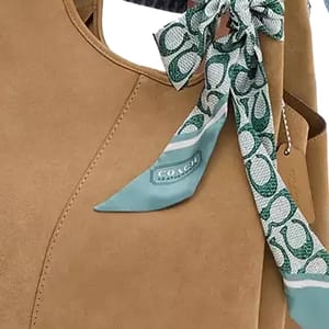 A silk skinny scarf with a green and white Coach signature print tied to a tan handbag.