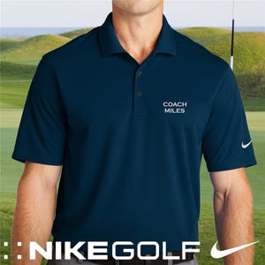 A blue Nike Golf polo shirt with \“Coach Miles\“ embroidered on the chest, featuring the Nike logo on the sleeve, set against a golf course background.