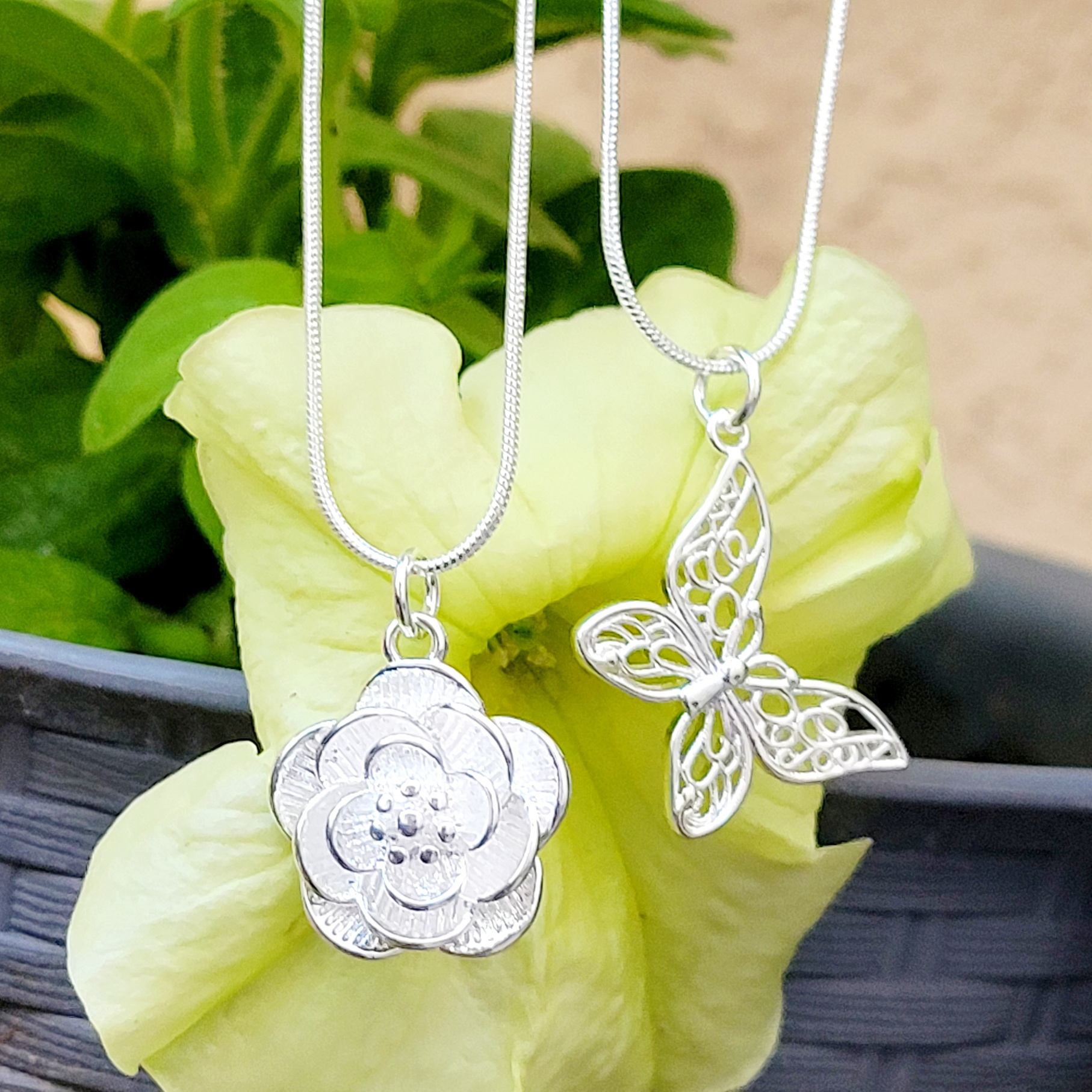 Silver necklaces with pendant charms, one featuring a flower design and the other a butterfly, displayed against a soft yellow flower background.