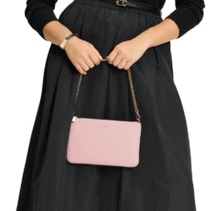 A person holds a Kate Spade Taylor wristlet with a pink leather body and gold chain strap, paired with a black skirt and sweater.