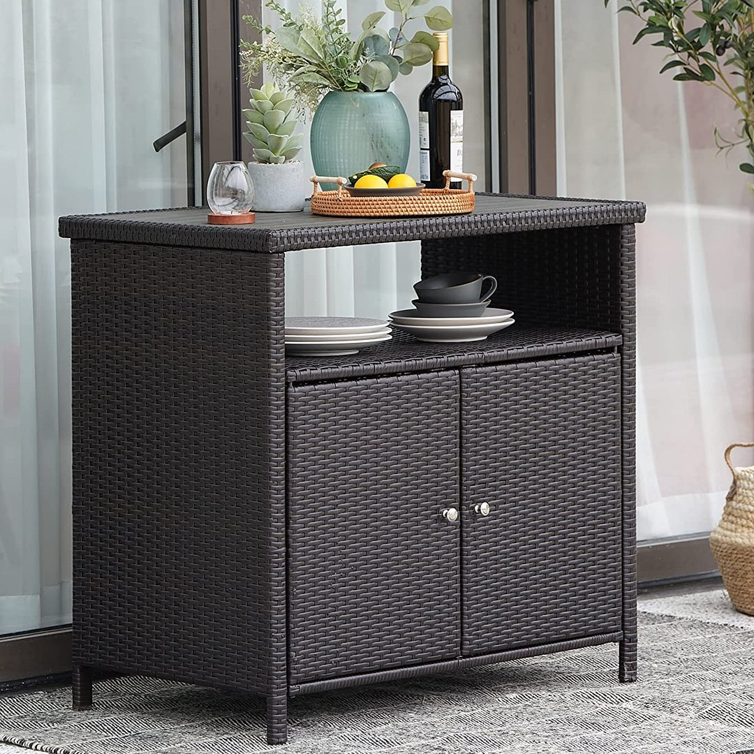 A dark wicker outdoor cabinet with an open shelf holding plates and cups, and a top surface displaying a tray with fruit, a vase, and a bottle of wine.