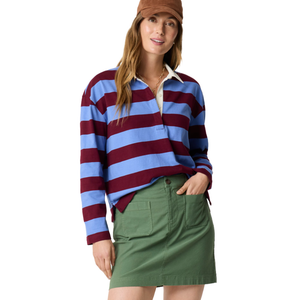 A woman wearing a striped rugby shirt in maroon and blue, paired with a green skirt featuring front pockets and a brown cap.