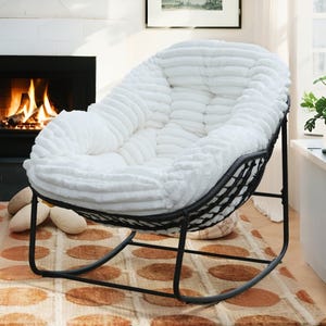 A plush, white rocking chair with a textured cushion sits on a patterned rug in front of a lit fireplace, creating a cozy atmosphere.